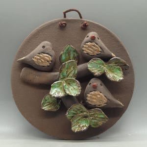 Può includere: Un ornamento da parete rotondo in ceramica marrone con quattro uccelli stilizzati appollaiati su un ramo con foglie verdi. Gli uccelli hanno il petto dorato e occhi rossi. Un cordino marrone è attaccato per appenderlo.