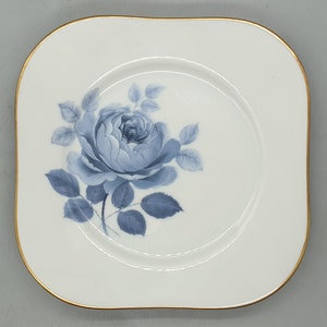 Puede incluir: Un plato cuadrado blanco con un borde dorado presenta un diseño floral azul y blanco de una sola rosa con hojas.