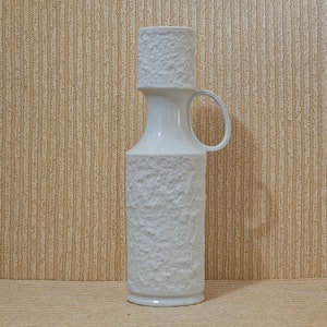 Könnte beinhalten: Weiße Keramikvase mit strukturierter Oberfläche und Henkel. Die Vase hat eine breite Öffnung und einen schmalen Hals.