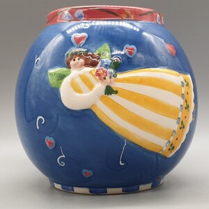 May include: A blue ceramic vase featuring a white and yellow striped angel design. The angel is holding a bouquet of flowers. The vase has a red and white rim and a blue and white base.