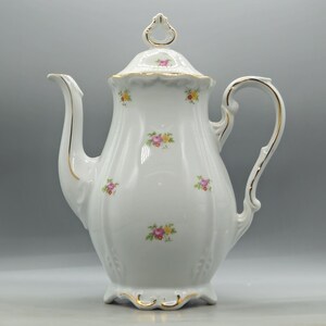 May include: A white porcelain teapot featuring a delicate floral pattern of small pink and yellow roses with green leaves. The teapot has a scalloped base, a curved handle, a long spout, and a lid with a finial, all accented with gold trim.