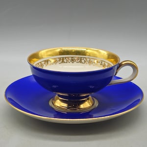 May include: A cobalt blue teacup and saucer set with gold trim. The teacup has a gold interior and handle. The saucer and the base of the teacup are also gold. The teacup has a decorative gold pattern around the rim.