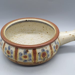 Può includere: Una zuppiera in ceramica con manico. La ciotola ha un interno ed esterno color crema con motivi floreali marroni e blu. Il bordo e il manico sono marroni.