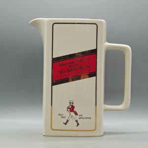 Puede incluir: Jarra rectangular de color crema con asa. La parte delantera presenta una etiqueta roja y dorada con el texto "Johnnie Walker Old Scotch Whisky". Debajo, un dibujo de un hombre con abrigo rojo. También es visible el texto "Born 1820 still going strong".