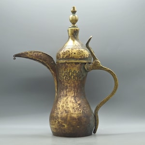 May include: An antique brass coffee pot with a long spout and curved handle. The pot has a decorative pattern near the top and a small finial on the lid. The brass has a tarnished, aged appearance, suggesting it is vintage.