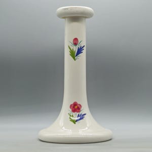 May include: A tall, off-white ceramic candlestick holder with a flared base and a rounded top. The candlestick is decorated with hand-painted floral designs in red, blue, and green. The overall design is simple and elegant.