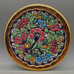 May include: A round, decorative plate with a gold rim and a colorful floral design. The plate features red, pink, blue, green, and yellow flowers and leaves on a white background. The rim has a dark blue border.