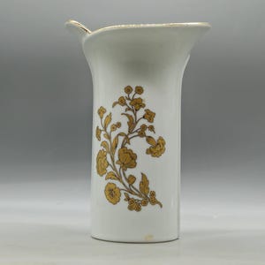 May include: A white ceramic vase with a gold floral design. The vase has a unique shape with a flared top and a small spout. The gold floral pattern is on the front of the vase.
