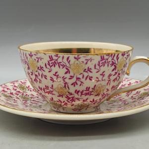 May include: A white teacup and saucer with a pink and gold floral pattern. The cup has a gold rim and a handle.