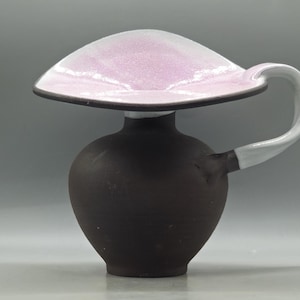 Puede incluir: Una jarra de cerámica única con una parte superior ancha esmaltada en rosa y un cuerpo marrón oscuro. El asa es blanca, curvándose elegantemente desde el lateral. El diseño es moderno y artístico, adecuado para servir bebidas o como pieza decorativa.