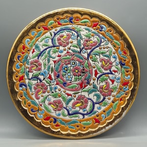 May include: Decorative ceramic plate with a gold rim and intricate floral design. The plate features a white background with pink, blue, green, and yellow flowers and leaves. The border is a detailed pattern of orange, red, and gold.