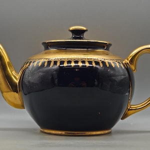 Op de afbeelding: Een zwarte en gouden theepot met een afgeronde body, een gebogen tuit en een lusvormige handgreep. De theepot heeft een gouden deksel en afwerking, met een decoratieve gouden band rond de bovenkant. De theepot is waarschijnlijk gemaakt van keramiek of porselein.