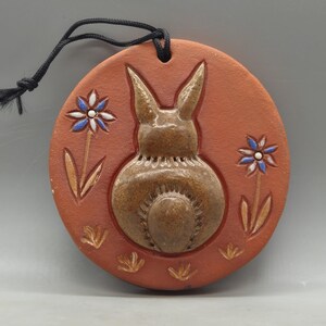 May include: A circular terracotta ceramic Easter decoration with a raised relief of a bunny and two stylized flowers with blue and white petals. It has a black cord for hanging.