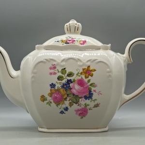 Könnte beinhalten: Cremefarbene Porzellan-Teekanne mit floralem Design mit rosa Rosen, blauen und gelben Blumen und grünen Blättern. Die Teekanne hat einen goldenen Rand an Griff, Ausguss und Deckel. Ein dekoratives Stück zum Servieren von Tee.