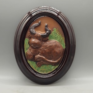May include: A brown ceramic wall plaque with a raised image of a brown bull with horns, lying down in a field of green grass. The plaque is framed in a dark brown oval frame.