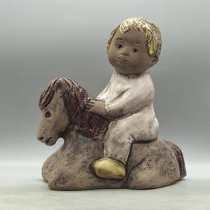 May include: A ceramic figurine of a child riding a rocking horse. The child is wearing a white onesie and has brown hair. The rocking horse is brown and has a brown mane and tail.