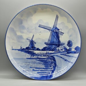 Könnte beinhalten: Großer, runder Keramikteller mit einer blau-weißen Windmühlenszene. Der Teller zeigt eine detaillierte Landschaft mit Windmühlen, Wasser und einem bewölkten Himmel. Der Rand ist dunkelblau.