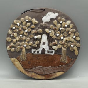May include: A circular ceramic wall plaque featuring a relief design of a house with a white roof and chimney smoke, flanked by two trees with textured brown trunks and light brown foliage with white accents, set against a dark brown background.
