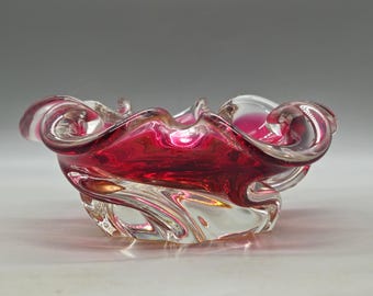 May include: A decorative glass bowl with a vibrant red interior and clear, swirling edges. The bowl has a unique, organic shape with ruffled rims, creating a visually appealing piece. The glass appears to be hand-blown, showcasing a blend of colors.
