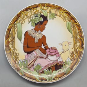 May include: Decorative plate with a seated figure and a lamb, wearing jewellery and a floral headpiece. The plate has a brown and green border and a cream background. The figure is wearing a pink and white patterned skirt.