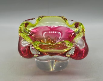 May include: A colorful glass ashtray with a unique shape. The ashtray features a combination of pink, yellow, and clear glass. The design is a mix of curves and angles, creating a visually interesting piece.