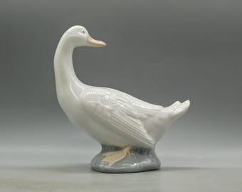 May include: A white porcelain figurine of a goose with a long neck and a detailed feathered body. The goose is standing on a gray base.