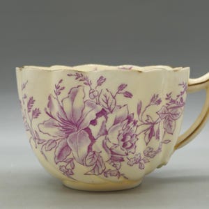 May include: A cream-colored ceramic teacup featuring a scalloped rim and a gold-toned handle. The exterior is decorated with a purple floral pattern of lilies and leaves. The cup has a fluted, ribbed body design with a smooth, glossy finish.