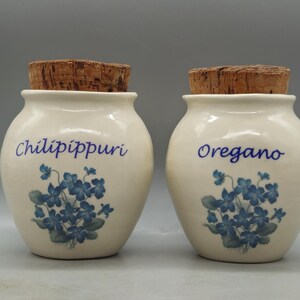 May include: Two white ceramic spice jars with cork lids. The jars have blue floral designs and the words "Chilipippuri" and "Oregano" printed on them.