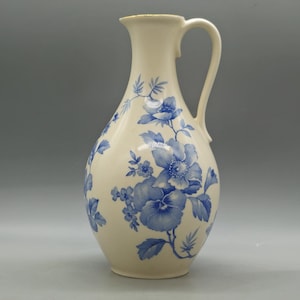 Op de afbeelding: Crèmekleurige keramische kan met een blauw bloemenontwerp. De kan heeft een slanke hals, een afgeronde body en een gebogen handvat. Het blauwe bloemenpatroon bevat grote bloemen en bladeren.