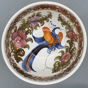 May include: A white ceramic bowl with a hand-painted design. The bowl features a central image of a bird with blue and orange plumage, surrounded by a floral border with pink and purple flowers and green leaves. The rim has a brown floral pattern.