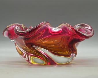 May include: A vibrant, hand-blown glass bowl with a ruffled edge. The bowl features a blend of colors, including shades of red, orange, and white. The glass has a glossy finish, and the design is swirled and textured.