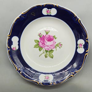 May include: A white porcelain plate with a navy blue rim and gold trim. The plate features a pink rose floral design in the centre and small floral accents on the rim. The plate has a scalloped edge.