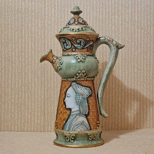 May include: A green and gold ceramic pitcher with a lid and a handle. The pitcher has a detailed design with a woman's face in profile on the front.