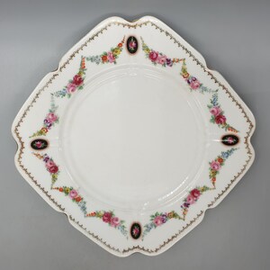 May include: A white square plate with a gold rim and floral design. The plate has a scalloped edge and features a repeating pattern of pink, red, and blue flowers with green leaves. The center of the plate is plain white.