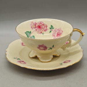 May include: Cream-colored teacup and saucer set with pink rose floral design and gold trim. The teacup has a pedestal base and a gold handle. The saucer has a scalloped edge and matching floral accents.