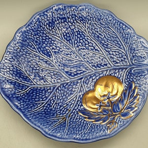 Könnte beinhalten: Eine blaue, keramik-gefertigte Servierplatte in Blattform mit einem erhabenen Design und goldenen Akzenten. Die Platte zeigt zwei goldfarbene Tomaten und Blätter.