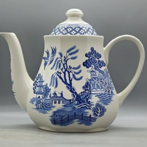 Può includere: Una teiera bianca con un motivo a salice blu e bianco. La teiera ha un coperchio con un motivo geometrico e un beccuccio sul lato sinistro. La teiera è decorata con una scena di una pagoda, un ponte e alberi.