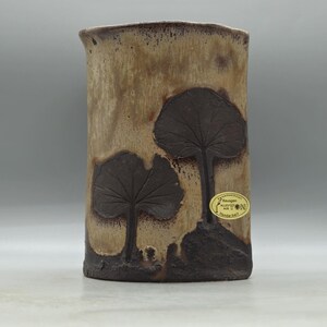 May include: A cylindrical ceramic vase with a textured, mottled tan and brown finish. Two dark brown leaf designs are embossed on the front. A small, round sticker with text is affixed to the side. The vase has a slightly irregular rim.