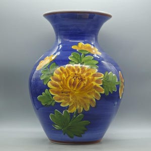 Op de afbeelding: Een blauwe keramische vaas met een brede basis en een uitlopende rand. De vaas is versierd met gele en oranje bloemmotieven en groene bladeren. De vaas heeft een witte rand en een bruine band aan de bovenkant.