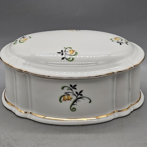 May include: A white porcelain trinket box with a lid. The box is decorated with a floral pattern in shades of green, yellow, and black. The box has a gold trim around the edge.