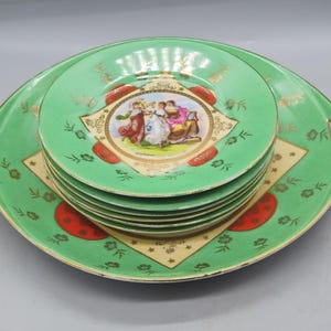 Könnte beinhalten: Ein Satz grüner Porzellanteller und ein Serviertablett. Die Teller und das Tablett weisen goldene Details und ein zentrales Design mit Figuren in historischer Kleidung auf. Das Tablett hat Griffe und rote Akzente.
