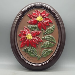 Può includere: Decorazione murale ovale con un rilievo di due fiori di poinsettia rossi con centri gialli e foglie verdi. I fiori sono posti su uno sfondo marrone all'interno di una cornice marrone scuro. La firma dell'artista è visibile.