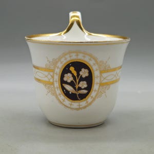 Könnte beinhalten: Eine weiße Zuckerdose aus Porzellan mit einem zentralen ovalen Medaillon auf dunkelblauem Grund und einem goldenen Blumenmotiv. Die Schale ist mit Goldverzierungen am Rand, am Henkel und an den Seiten sowie mit goldenen Filigrandetails versehen.