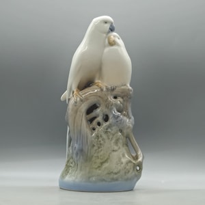 Peut inclure: Une sculpture en céramique blanche représentant deux oiseaux perchés sur une branche d'arbre. Les oiseaux sont proches l'un de l'autre, l'un d'eux niché contre l'autre. La base est de couleur bleu clair.
