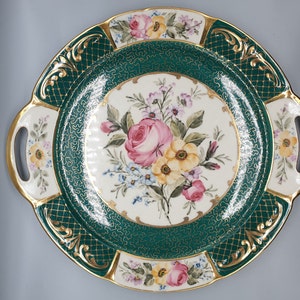 May include: A round serving platter with two handles, featuring a green and gold design with a floral motif. The center of the platter has a white background with a bouquet of pink, yellow, and white flowers.