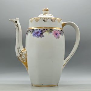 May include: A white porcelain coffee pot featuring a floral band with purple and pink flowers around the upper body. The pot includes a lid with a gold-trimmed knob, a curved handle, and a spout decorated with gold filigree and floral accents.