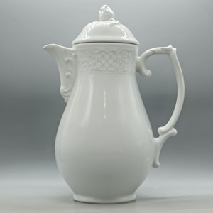 May include: A white porcelain coffee pot featuring a smooth, glossy finish, a curved handle, and a spout with decorative scrollwork. The upper section includes a textured lattice pattern, and the lid is topped with a small, sculpted finial.