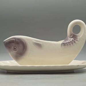 May include: A cream-coloured ceramic gravy boat and matching saucer, shaped like a fish. The fish-shaped boat has a purple eye and fin details, with a loop handle. The saucer is oval-shaped.