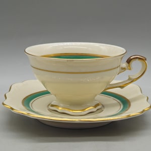 Puede incluir: Juego de taza y platillo de color crema con ribete dorado y una banda verde. La taza tiene un asa curvada y se apoya en un platillo a juego con un borde festoneado. Un juego de té clásico.