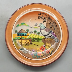 May include: A ceramic plate featuring a hand-painted design of a bird, a small house, and a wagon. The plate has a brown rim and a white centre with a colourful scene.